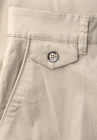 Nærbillede af beige bukselomme med klap, der er sikret med en firehullet knap, og et bæltestrop over lommen.