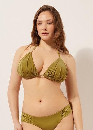 Ung kvinde med bølget brunt hår iført olivengrøn bikini med rynkede detaljer på top og matchende bund foran en lys baggrund.