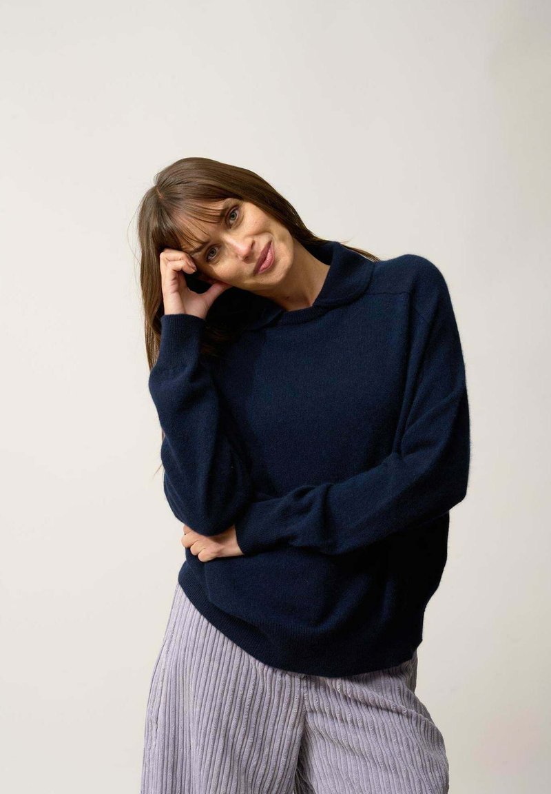 Granatowy sweter z wysokim dekoltem, luźny krój i prążkowane mankiety, w połączeniu z jasnymi, teksturowanymi spodniami o szerokich nogawkach.
