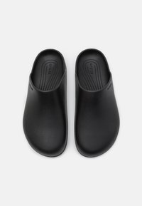 Scarpe slip-on nere realizzate in materiale liscio, con forma arrotondata, design minimale e logo in rilievo sul lato.