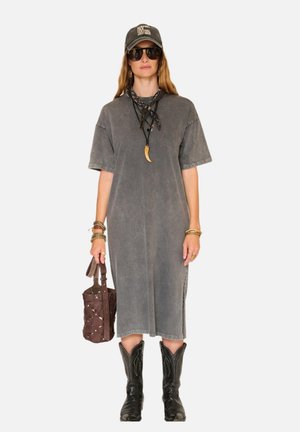 Femme portant une robe midi grise délavée, des bottes de cowboy noires, une casquette noire, des lunettes de soleil foncées, des colliers superposés, des bracelets, et tenant un sac à main marron.