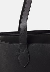 Musta kassi, jossa on teksturoitu pinta, varustettu kahdella pitkällä kahvalla ja kaarevalla yksityiskohdalla hihnassa, korostaen minimalistista muotoilua.