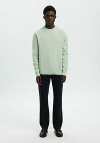 Młody mężczyzna ubrany w jasnoszary sweter z dzianiny, ciemne jeansy o prostym kroju i czarne skórzane buty, stojący na jednolitym jasnym tle.