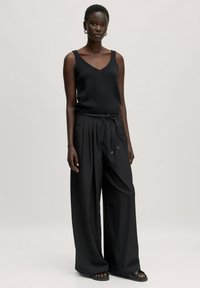 Schwarzes, gestricktes Tanktop mit V-Ausschnitt, kombiniert mit lockeren, schwarzen, gefalteten Hosen mit einem Kordelzugbund. Flache, schwarze Sandalen runden den Look ab.