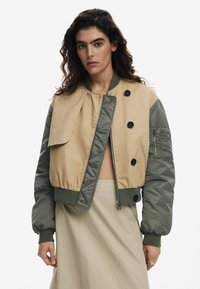 Tan and green bomber jacket with a cropped design, featuring a button closure, oversized collar, and zipped pocket on the sleeve.