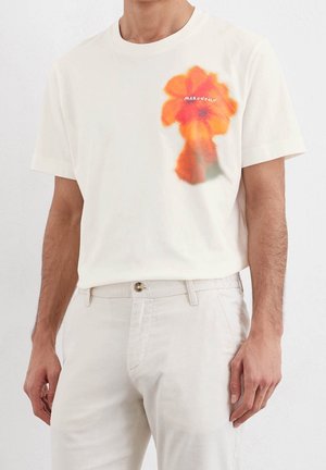 Man wearing white short-sleeve t-shirt and pants, shirt features blurred orange flower and small white text on chest.