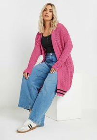 Rosa stickad cardigan med lös passform, öppen front och intrikat mönster; kombinerad med högmidjade blå jeans och vita sneakers med svarta ränder.