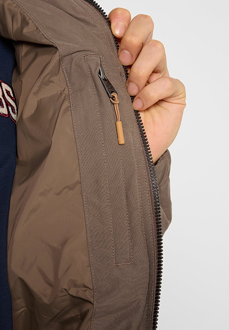 Giacca bomber marrone con una texture liscia e lucida. Presenta una tasca con cerniera sulla manica e una zip con una linguetta marrone chiaro.