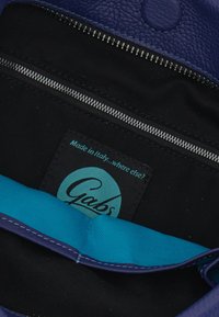 Interno in pelle viola di una borsa con rivestimento nero, dotato di una tasca con cerniera e un accento in tessuto blu con un'etichetta rotonda con il logo Gabs.
