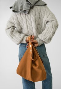 Borsa di stoffa beige con forma morbida, texture liscia e dettagli metallici sulla maniglia; tenuta da una persona che indossa un maglione grigio a trecce.