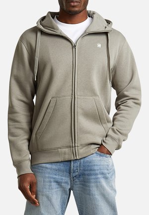 Man draagt een lichtgrijze hoodie met ritssluiting, voorzakken en koordjes, gecombineerd met lichtblauwe spijkerbroek en een wit T-shirt eronder.