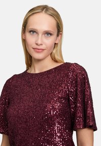 Top bordeaux con paillettes, maniche corte e scollatura tonda, caratterizzato da una superficie testurizzata che riflette la luce.