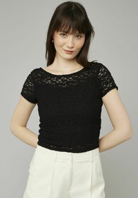 Haut noir à manches courtes en tissu texturé avec un motif floral et une silhouette ajustée, associé à un pantalon de couleur claire.