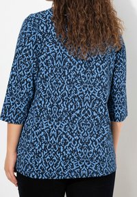 Blå mönstrad topp med trekvartsärmar, med en mörkbå bas och ljusblå abstrakta mönster, tillverkad av ett texturerat tyg.