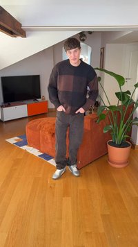 Fekete és barna foltos kötött pulóver, sötét farmer és ezüst sportcipő. Egy szobában áll, ahol nagy zöld növény és narancssárga kanapé van.