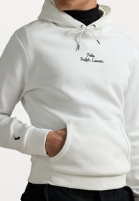 Vit hoodie i bomull med framficka, dragsko-kapuschong, broderad logga i svart och en liten logga på ärmen.