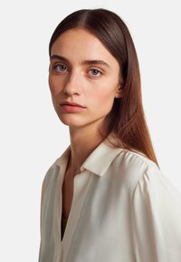 Blusa di crema chiaro con colletto e maniche leggermente arricciate, su uno sfondo neutro. Tessuto liscio e leggero.