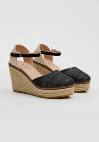 Sandalias de cuña negras con parte superior de tejido texturizado, tira de tobillo beige en contraste y plataforma estilo cuerda. Forro interior suave y costuras decorativas.