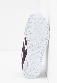 Semelle blanche de chaussure Reebok avec des motifs de grip texturés et un logo, positionnée verticalement contre un mur blanc d'angle.