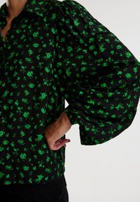 Camicia nera con motivo floreale verde, dotata di maniche a sbuffo e colletto con bottoni. Il tessuto sembra leggero con una leggera lucentezza.