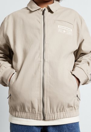 Beżowa kurtka na zamek z logo „Converse All Star” na piersi, ściągacze na mankietach, boczne kieszenie na zamek, noszona na białej koszuli i niebieskich dżinsach.