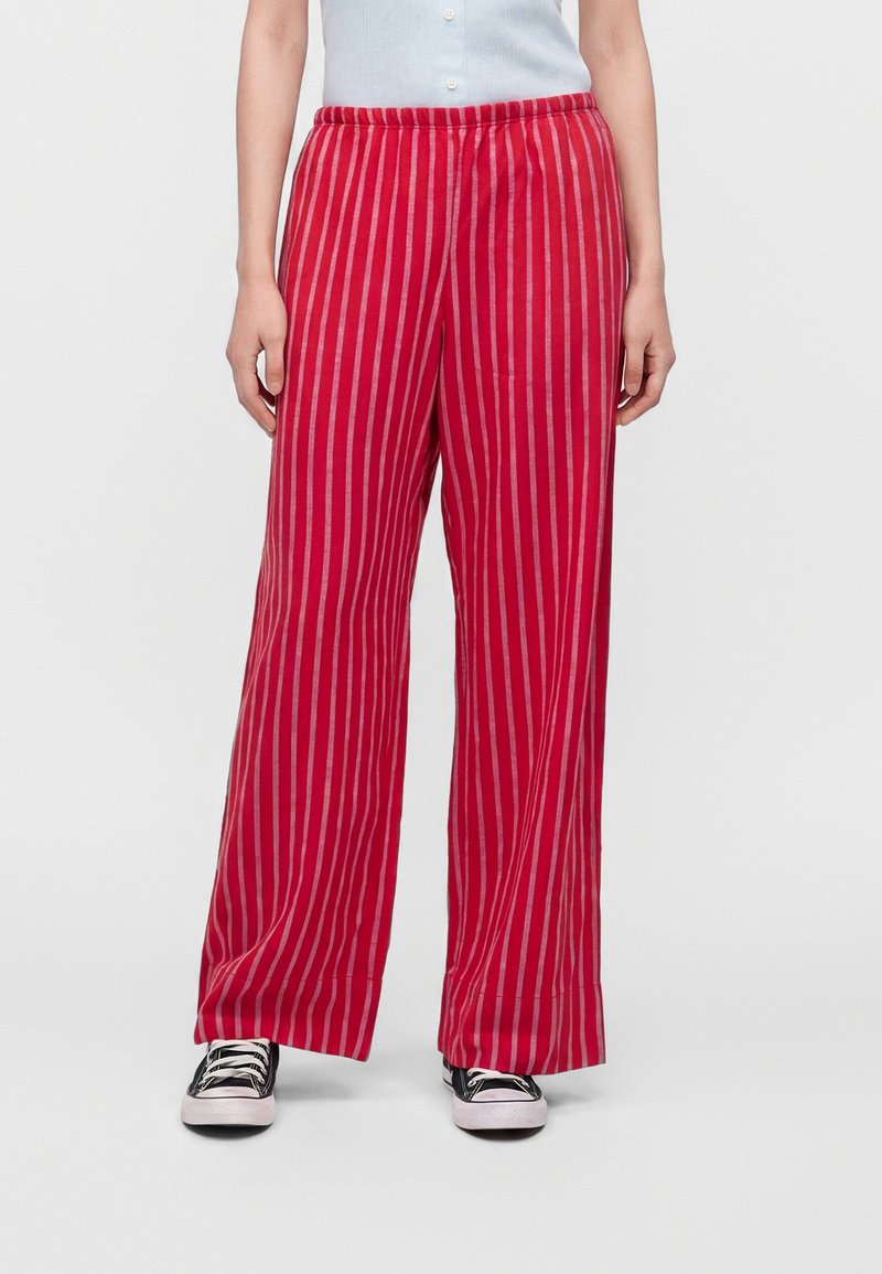 Personne portant un pantalon rouge ample à rayures verticales roses, des baskets noires et un haut sans manches bleu clair, debout devant un fond uni.