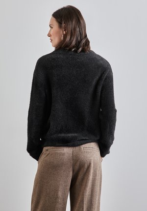 Maglione nero morbido con vestibilità comoda, abbinato a pantaloni marroni con texture. L'outfit ha dettagli minimi e un aspetto casual.