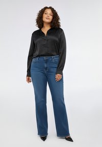 Camicia di raso nero con piccoli accenti metallici, maniche lunghe e chiusura frontale con bottoni, abbinata a jeans blu svasati e tacchi neri appuntiti.