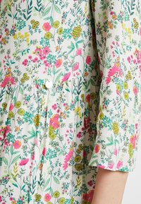 Robe florale avec un fond blanc parsemé de fleurs colorées roses, jaunes et vertes. Détail de boutons à la taille ; tissu doux et texturé.