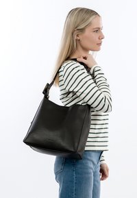 Blonde vrouw draagt een gestreept shirt met lange mouwen en een spijkerbroek en heeft een grote zwarte leren schoudertas met verstelbare riem bij zich.