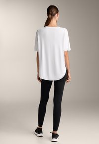 Bílý top s krátkým rukávem a kulatým spodním lemem, kombinovaný s černými legínami a černými sportovními botami, na neutrálním pozadí.
