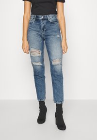 Höga blå denimjeans med slitna knäskärningar, rak passform och bältesöglor, i kombination med svarta ankelskor.