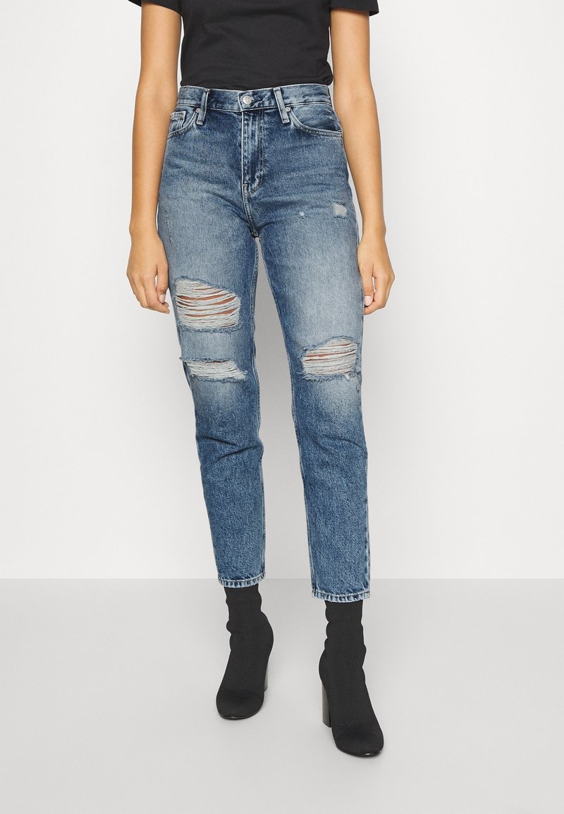 Höga blå denimjeans med slitna knäskärningar, rak passform och bältesöglor, i kombination med svarta ankelskor.