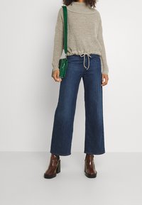 Vaaleanharmaa neulottu paita, jossa on käännetty kaulus, yhdistetty tummansinisiin leveälahkeisiin farkkuihin. Asun viimeistelee vihreä olkalaukku ja ruskeat korolliset saappaat.