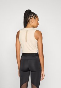 Béžový tielkový top s výstrihom na chrbte, v kombinácii s čiernymi legínami s vysokým pásom, ktoré majú sieťované detaily a hladký pás.