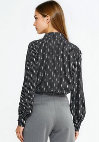 Czarna i biała bluzka z długim rękawem w pionowe paski i ozdobny wzór. Posiada wysoki kołnierz i marszczone rękawy, zestawiona z szarymi spodniami.