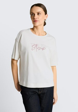 Frau mit zurückgebundenem Haar trägt ein weißes Kurzarmshirt mit dem Text „Time to Blossom“ und dunkle Jeans, steht vor einem einfarbigen hellen Hintergrund.
