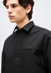 Νέος άνδρας που φοράει μαύρο πουκάμισο με κουμπιά, με διάτρητη τσέπη στο στήθος και το λογότυπο "BOSS" σε ανοιχτό γκρι φόντο.