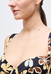 Femme portant une robe noire à fleurs beiges et brunes, une boucle d'oreille goutte dorée, et les cheveux attachés en queue de cheval.