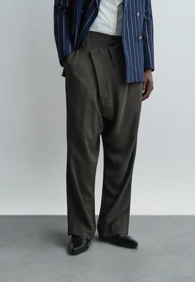 Personne portant un pantalon ample marron foncé, des chaussures noires à bouts pointus, une chemise blanche et un blazer bleu à rayures verticales jaunes.