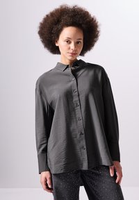 Camicia grigia scura con finitura testurizzata, maniche lunghe e colletto. Abbinata a jeans scuri con dettagli metallici.