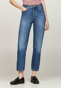 Visok pas modri denim jeans z ravnim krojem, ki imajo pet žepov in rahel učinek bledenja. Obuti so s črnimi loaferji.