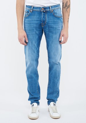 Uomo con jeans dritti azzurri chiari, sneakers bianche e camicia bianca; tatuaggio visibile sul braccio sinistro.