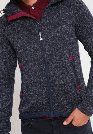 Mann trägt eine dunkel gesprenkelte Jacke mit Reißverschluss und roten Reißverschlussakzenten sowie ein bordeauxfarbenes Poloshirt darunter.