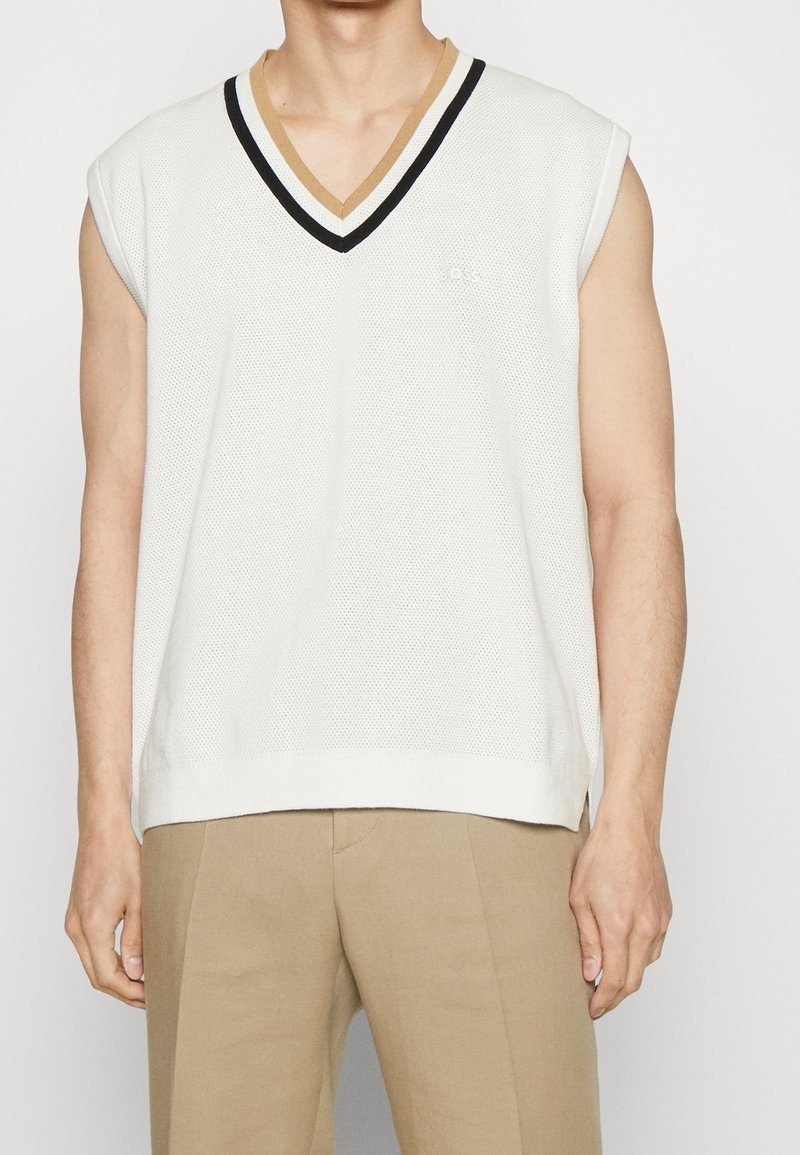 Gilet en maille blanche sans manches avec col en V orné de rayures noires et marron, porté sur un pantalon beige par une personne debout.