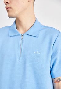 Pessoa jovem a usar uma camisa polo Adidas azul clara com um fecho na frente e a mostrar uma tatuagem colorida no braço.