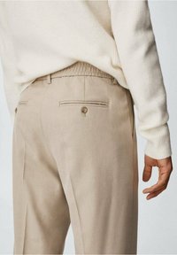 Pantaloni beige con una texture liscia, dotati di due tasche posteriori con bottoni e una sottile vita elasticizzata. Indossati con un maglione color crema.