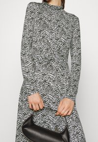 Eine Frau trägt ein langärmliges, schwarz-weiß gestreiftes Zebra-Kleid und hält eine kleine schwarze Handtasche an ihrem Tragegurt vor sich.