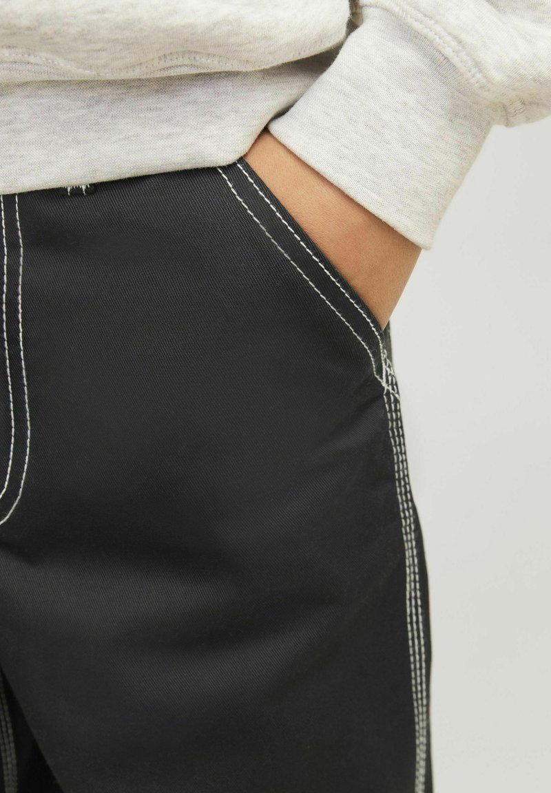 Svarta shorts med vit kontrastsömn, med en sidoficka och en texturerad tyg. En ärm av en grå sweatshirt syns ovanför.