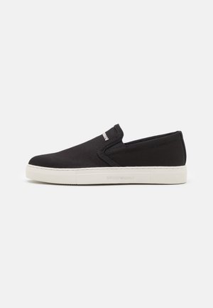 Musta slip-on kankaasta valmistettu tennari, jossa valkoinen kumipohja ja "Emporio Armani"-brändäys sivulla ja kielen päällä.